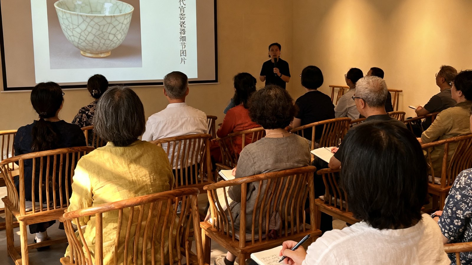 艺术美学沙龙现场，宽敞明亮的空间中投影幕布上展示着宋代官窑瓷器的细节图片，二十余位不同年龄的参与者坐在木质椅子上专注聆听，有人做着笔记